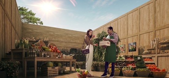 Mercearia repleta de vegetais com um comerciante com uma caixa de tomates e uma mulher a olhar para os tomates