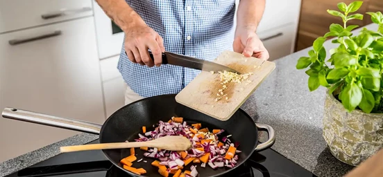 Homem coloca alho cortado na frideira que está a cozinhar os alimentos numa placa 