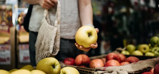 Maçã na mão de uma mulher em destaque rodeada de mais frutas num mercado 
